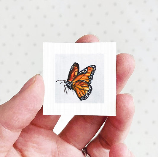 A hand holding a small square piece of paper with a colorful butterfly image on it.