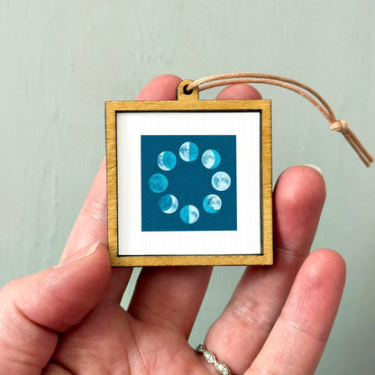 A hand holding a small wooden frame containing a framed image of the phases of the moon.