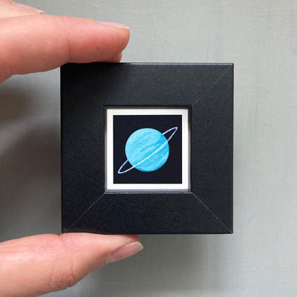 A hand is holding a small black square box with a white border, containing a small illustration of a blue planet with rings.