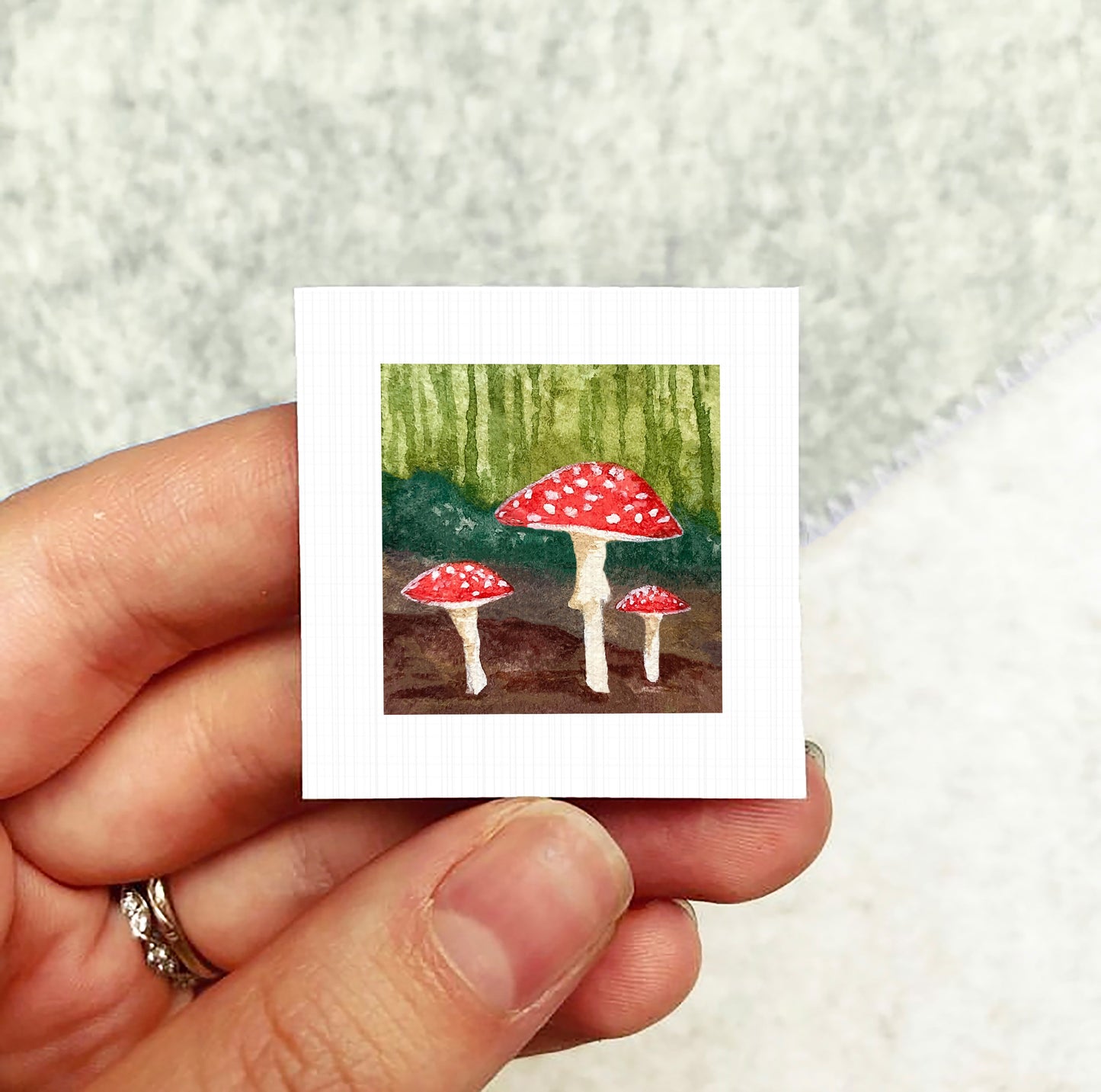A hand holding a small square card with a painting of three red mushrooms against a green background.