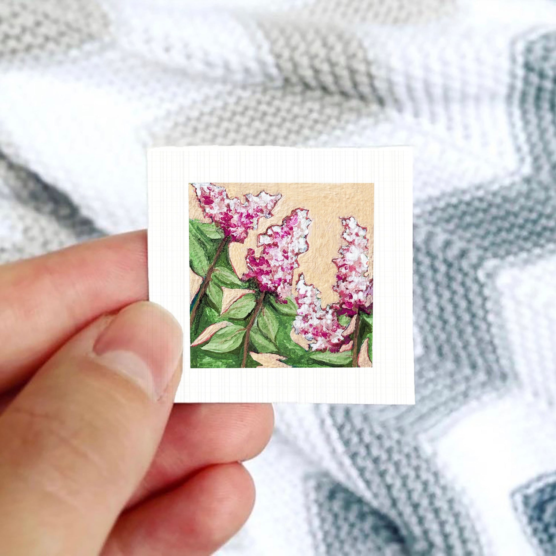 A hand is holding a small square card with a painting of pink flowers on it.