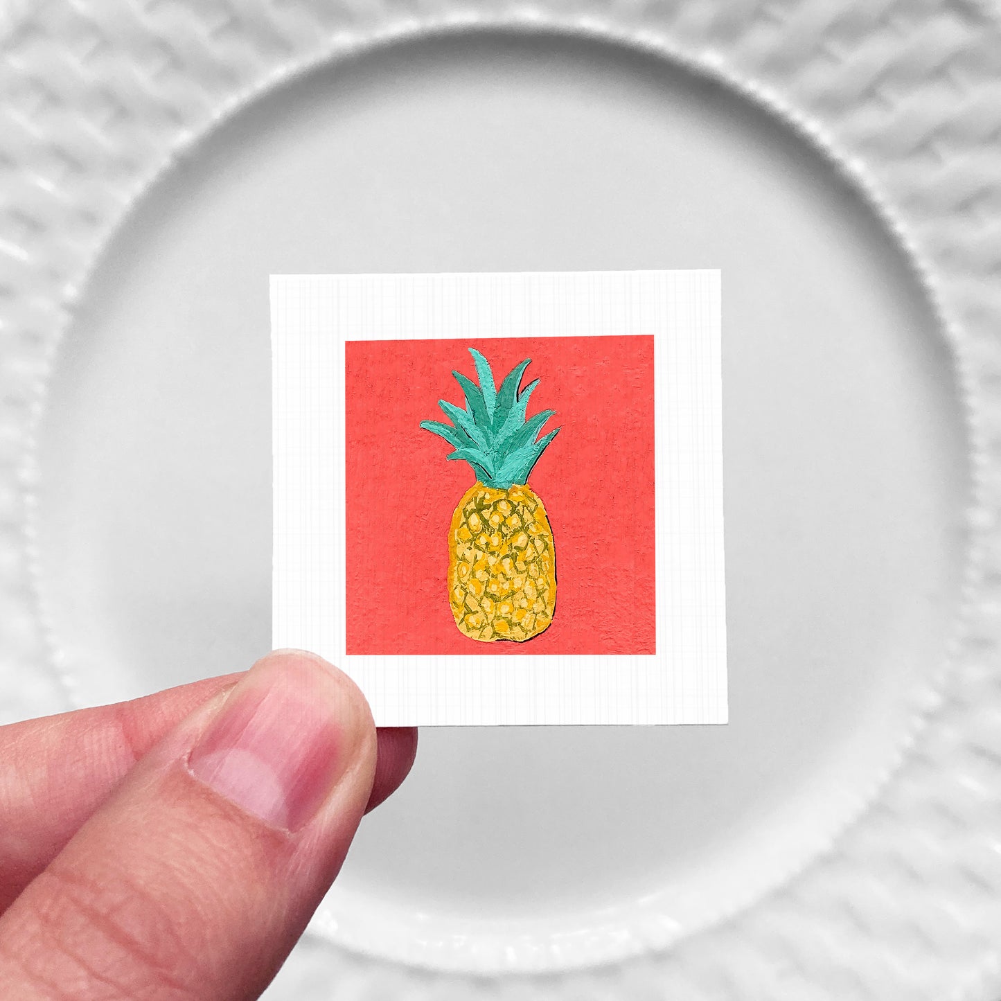A hand is holding a small square with a vibrant red background and a pineapple illustration on it.