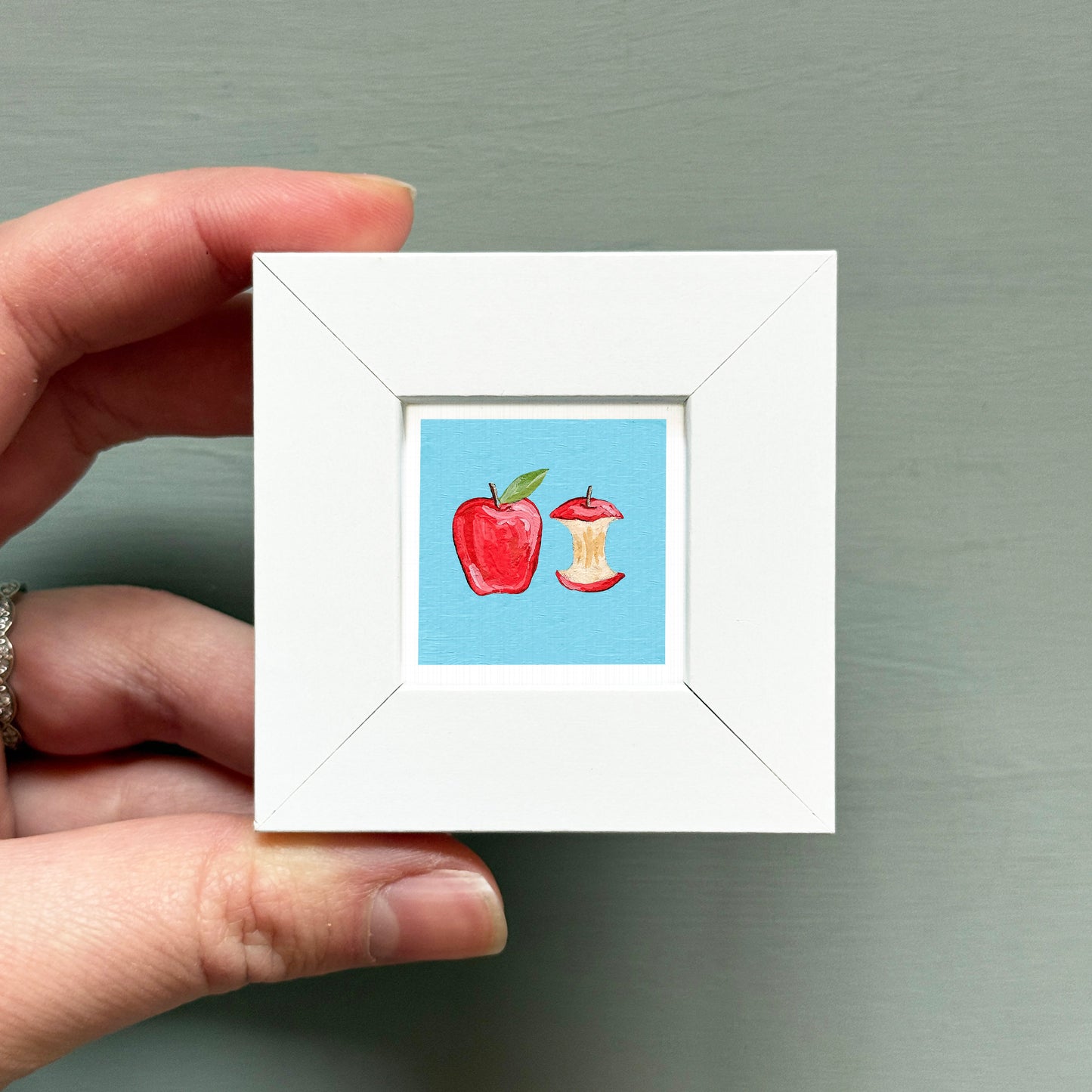 A hand is holding a small white square frame containing an illustration of an apple and an apple core.
