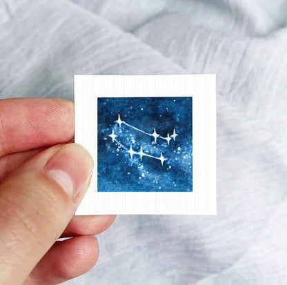 A hand is holding a small square-shaped paper with a depiction of a starry night sky on it.