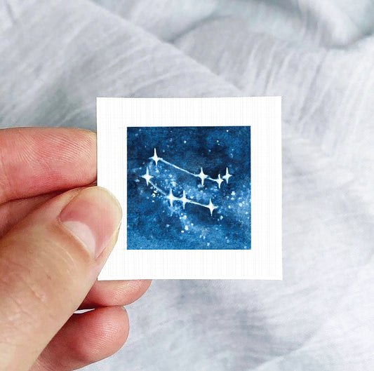 A hand is holding a small square-shaped paper with a depiction of a starry night sky on it.