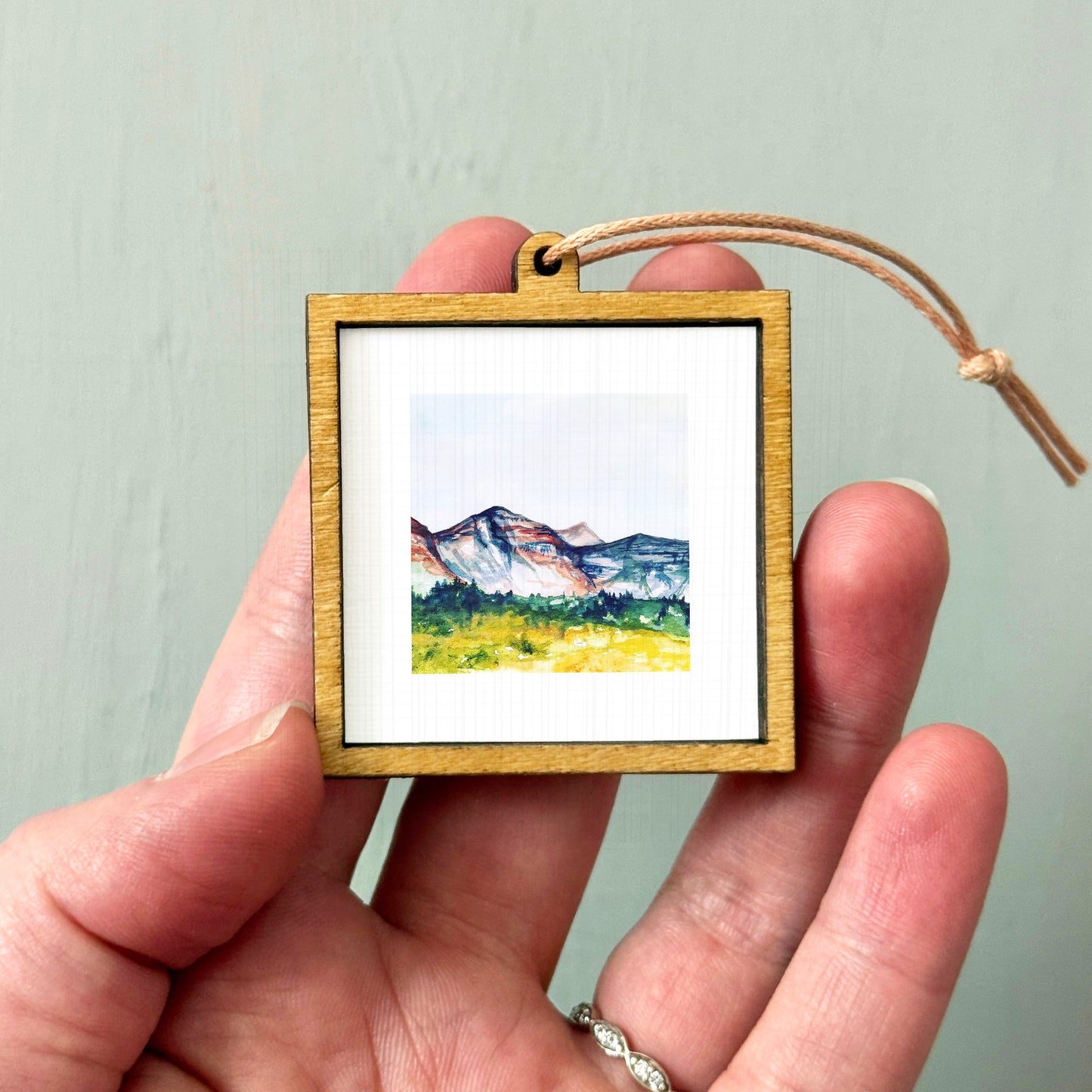 A hand holding a small wooden frame containing a watercolor painting of a mountain landscape.
