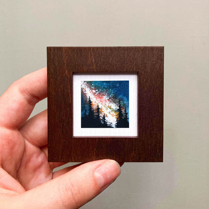 A hand is holding a small wooden frame containing a painting of a night sky with stars and silhouettes of trees.