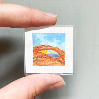 A person's hand holding a small square picture of a colorful landscape with mountains and a rainbow.