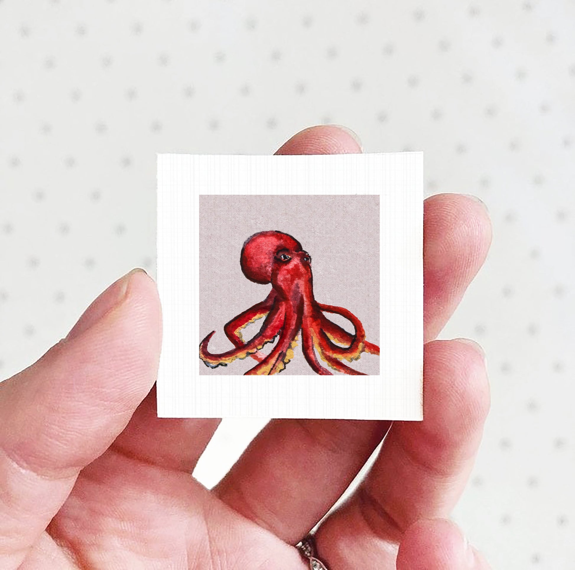 A hand holding a small square picture of a red octopus against a white background.