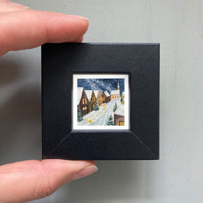 A hand is holding a small, square-shaped black frame containing a miniature painting of a snowy village scene.