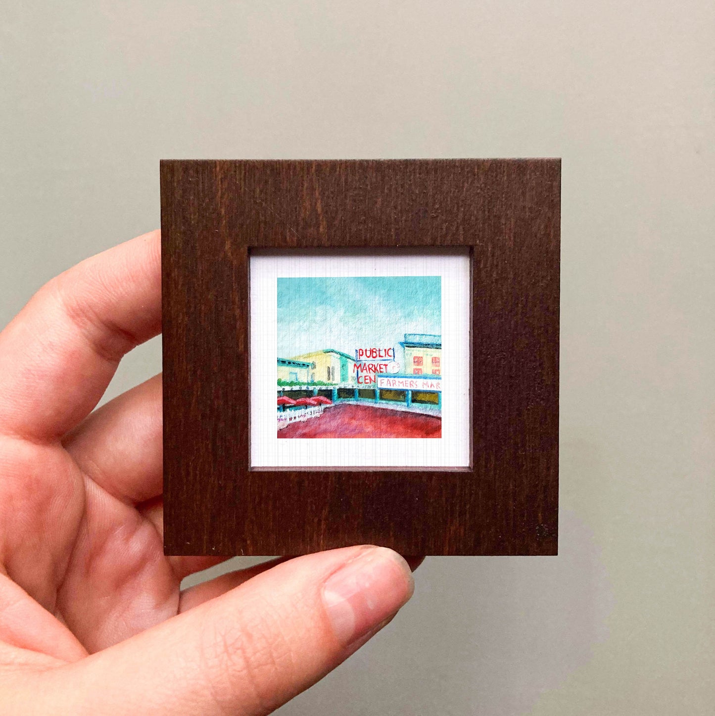 A hand is holding a small, square-shaped picture frame containing a colorful painting of a building.