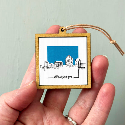 A hand holding a small wooden frame with a picture of a city skyline and the word "AlBUQUERQUE" written below it.