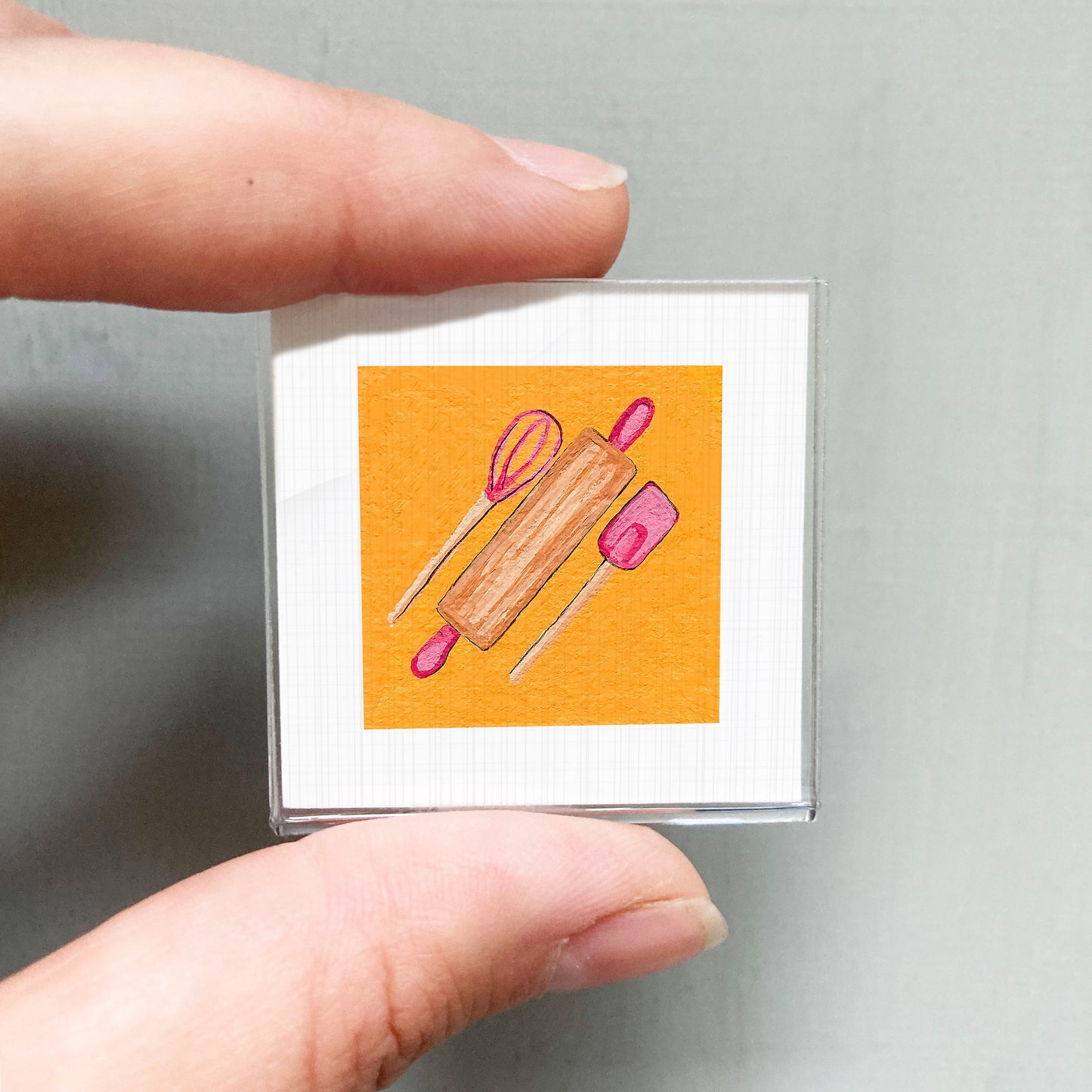 A person's hand holding a small square with an illustration of a kitchen utensil, such as a rolling pin or spatula, on an orange background.