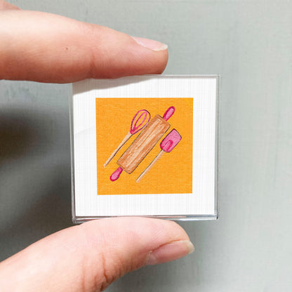 A person's hand holding a small square with an illustration of a kitchen utensil, such as a rolling pin or spatula, on an orange background.