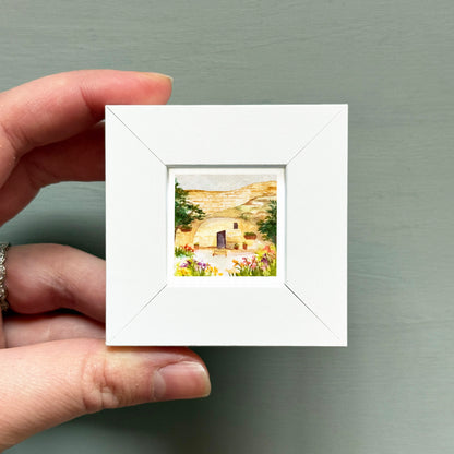A hand is holding a small white square frame containing a watercolor painting of a rural landscape with a house and flowers.