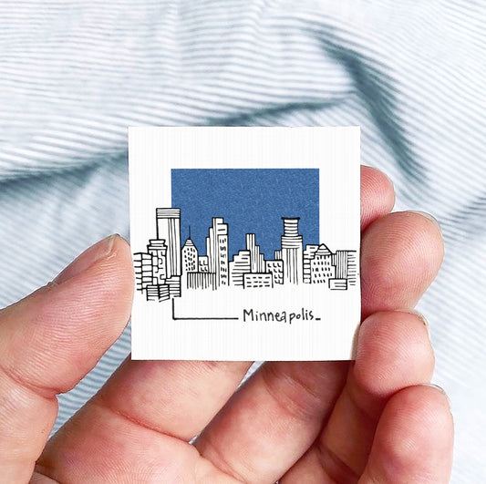 A hand holding a small square-shaped object with a drawing of a city skyline and the text "Minneapolis" written on it.