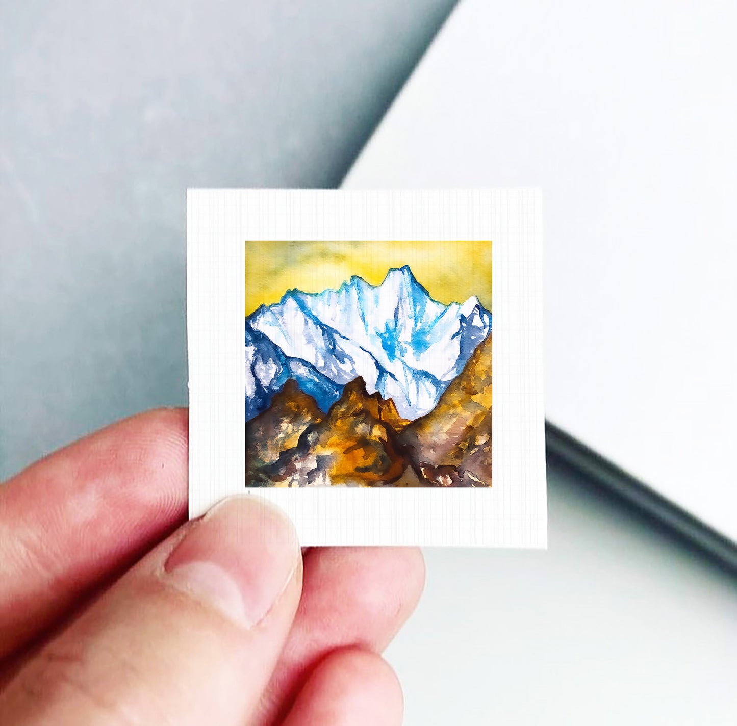 A hand is holding a small square-shaped piece of paper with a painting of a mountain landscape on it.