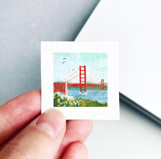 A person's hand holding a small square picture depicting the Golden Gate Bridge in San Francisco, California.