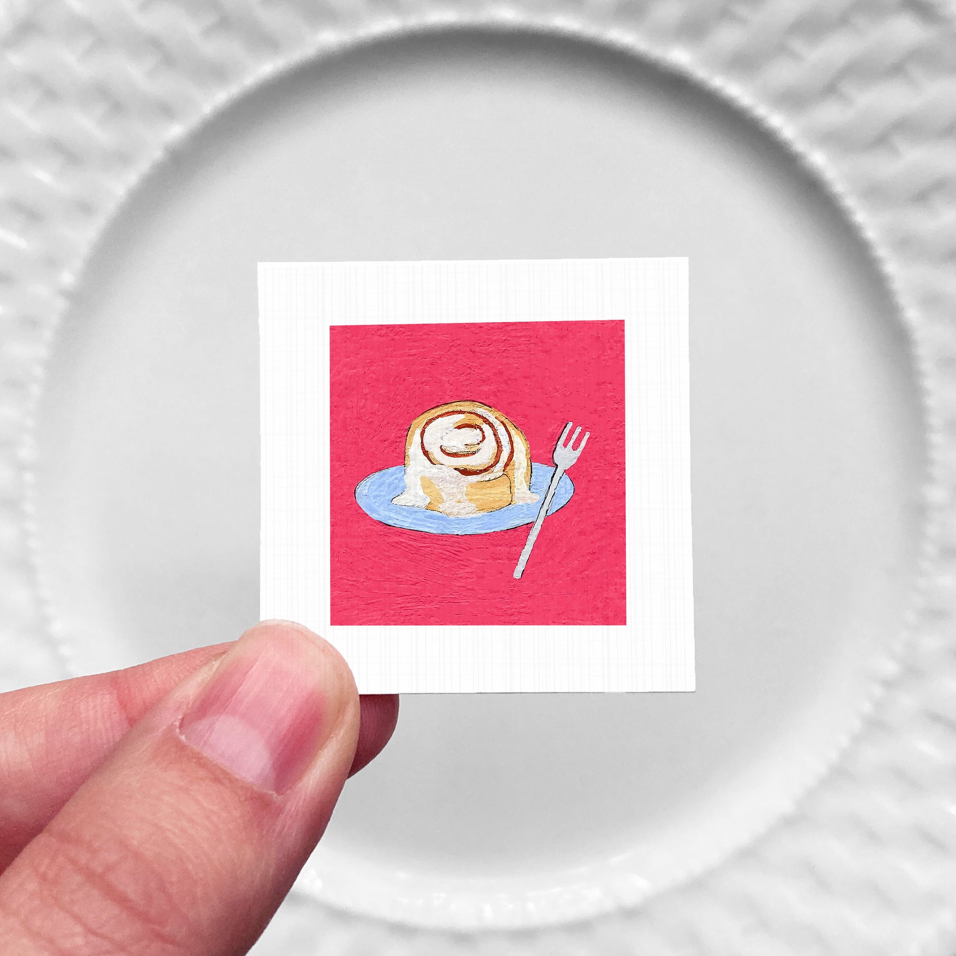 A hand is holding a small square card with an illustration of a cinnamon roll on a blue plate, against a white background.