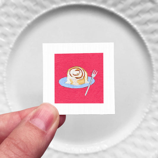 A hand is holding a small square card with an illustration of a cinnamon roll on a blue plate, against a white background.