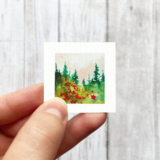 A hand is holding a small square painting of a forest scene with green trees and red flowers.