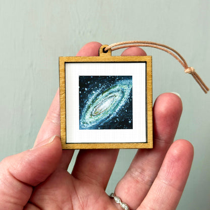 A hand holding a small wooden frame containing a framed image of a galaxy.