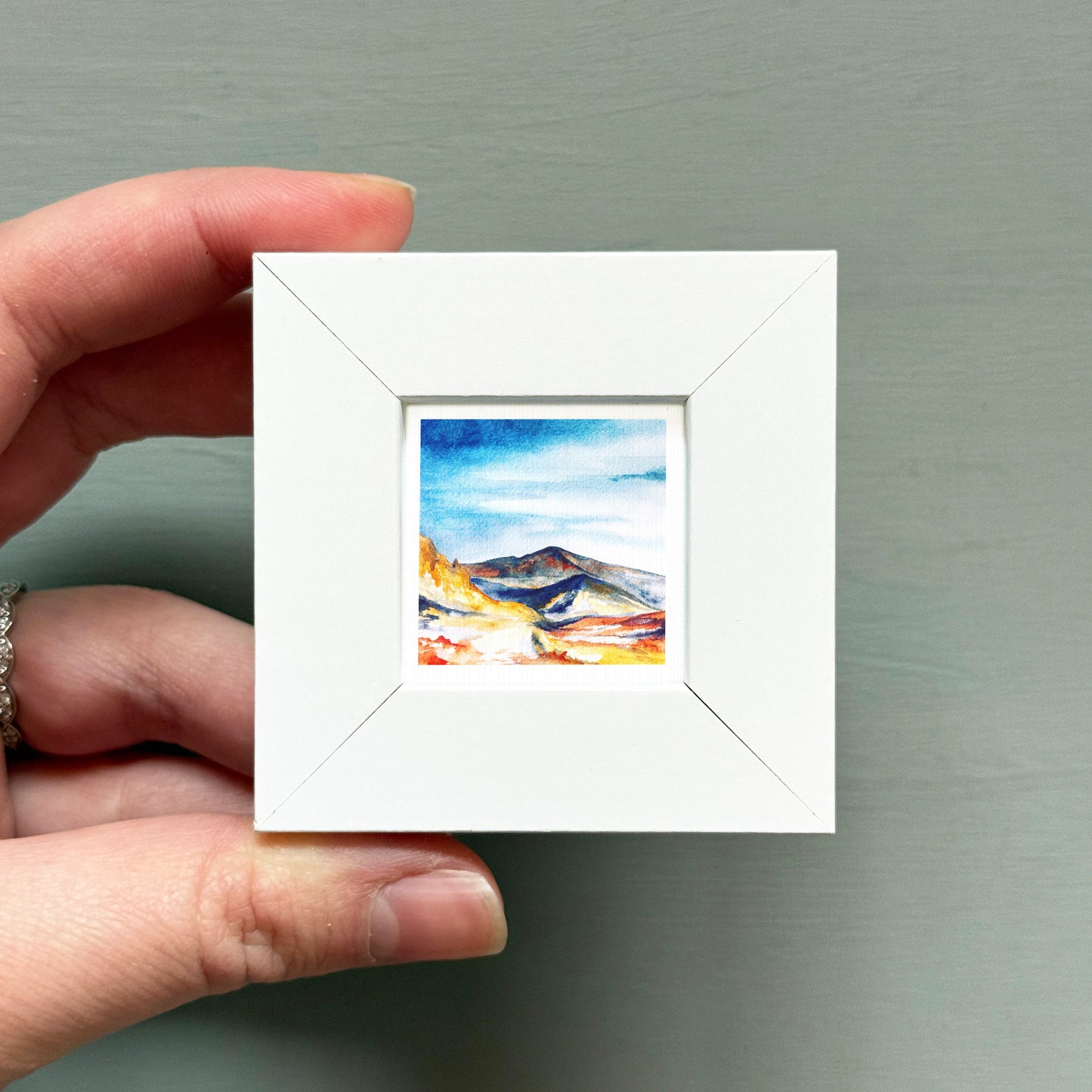 A hand is holding a small white square frame containing a watercolor painting of a mountain landscape.