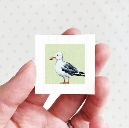 A hand holding a small square picture of a seagull against a green background.
