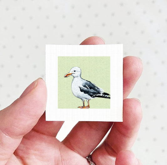 A hand holding a small square picture of a seagull against a green background.