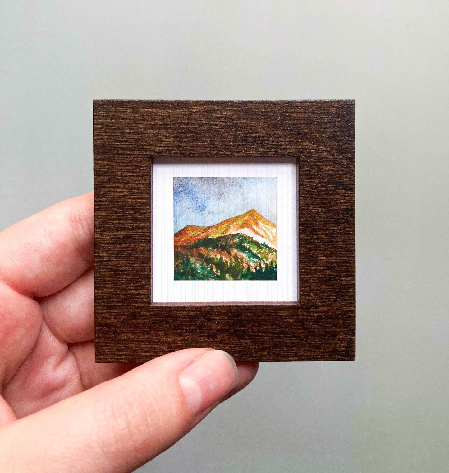 A hand is holding a small, square-shaped wooden frame containing a watercolor painting of a mountain landscape.