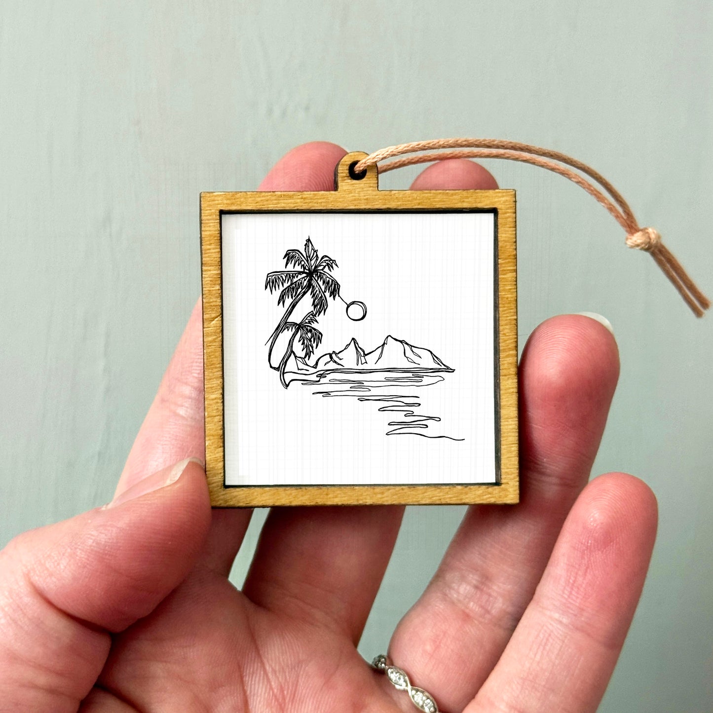 A hand holding a small wooden frame containing a black and white line drawing of a palm tree, mountains, and a body of water.