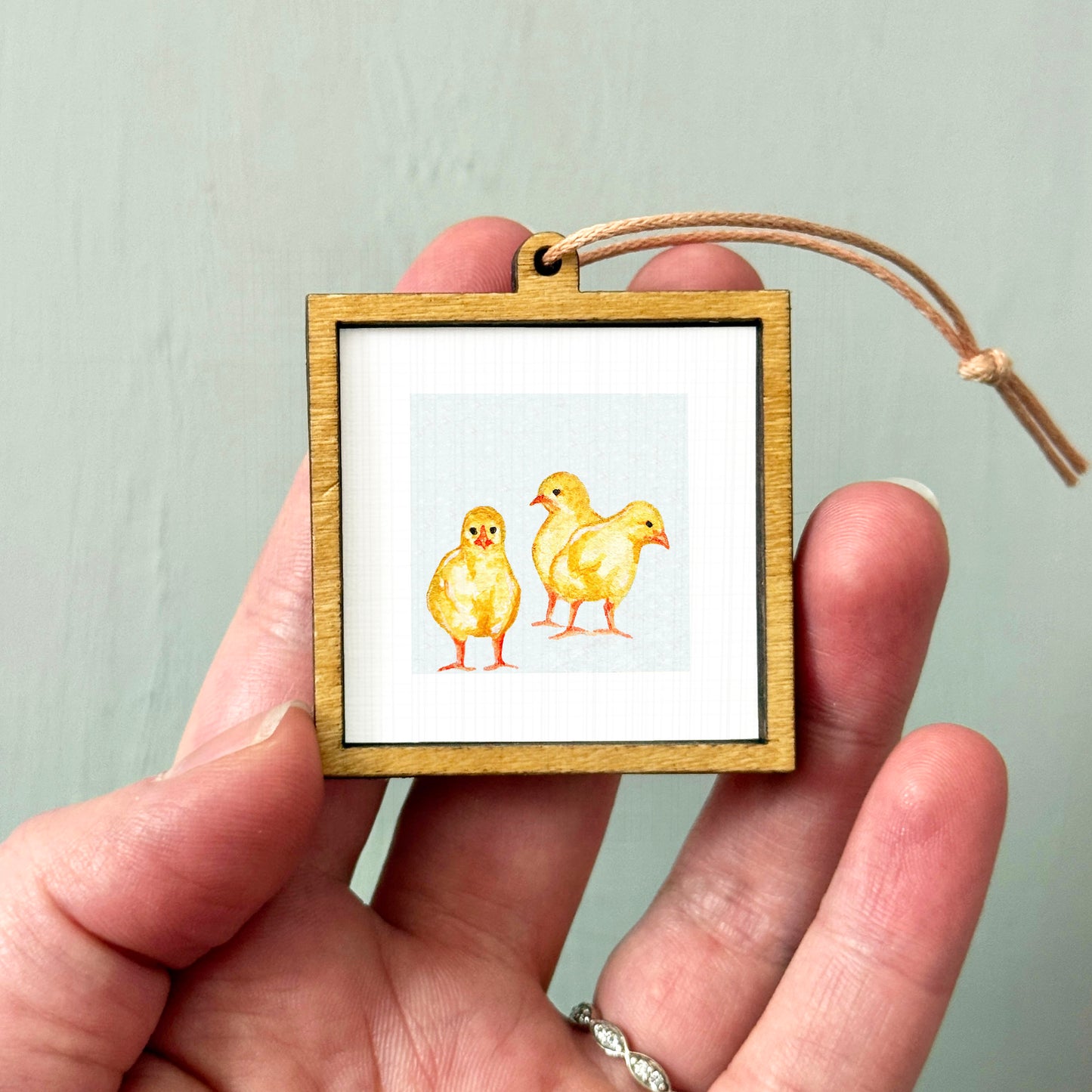 A person's hand holding a small wooden frame containing a picture of three yellow chicks.