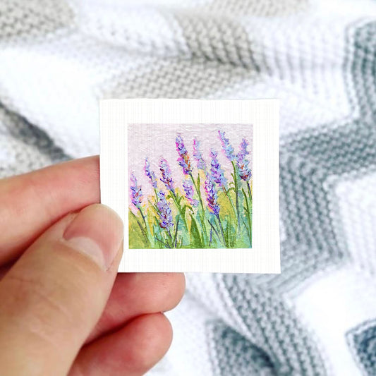 A hand is holding a small square card with a painting of purple flowers on it, against a background of a white and gray striped fabric.