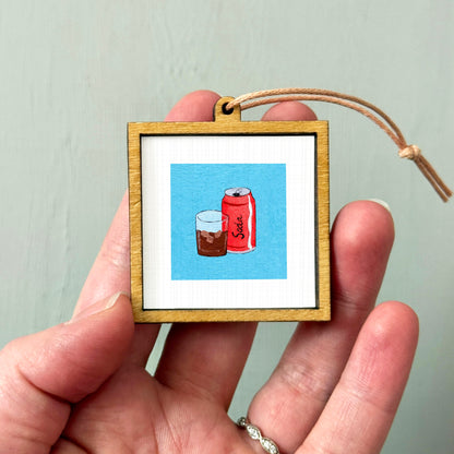 A person's hand holding a small wooden frame containing an illustration of a glass of soda and a can of soda.