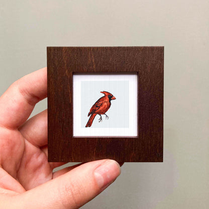A hand is holding a small wooden frame containing a framed image of a red cardinal bird.