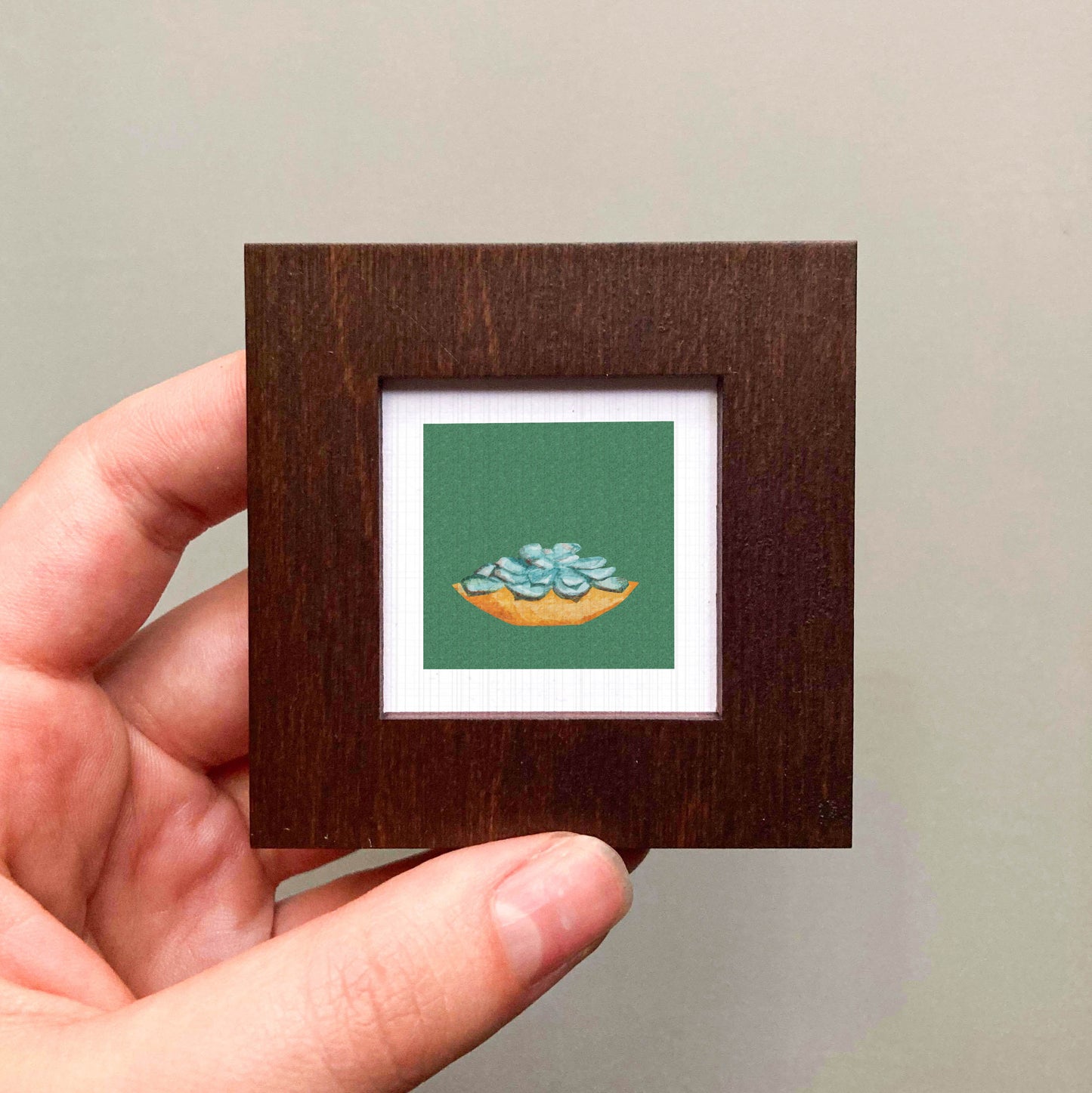 A hand is holding a small wooden frame containing a framed image of a green plant with blue flowers.