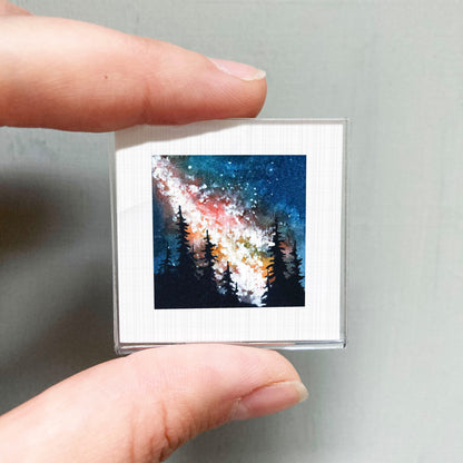 A hand is holding a small square picture of a night sky with trees in the background.
