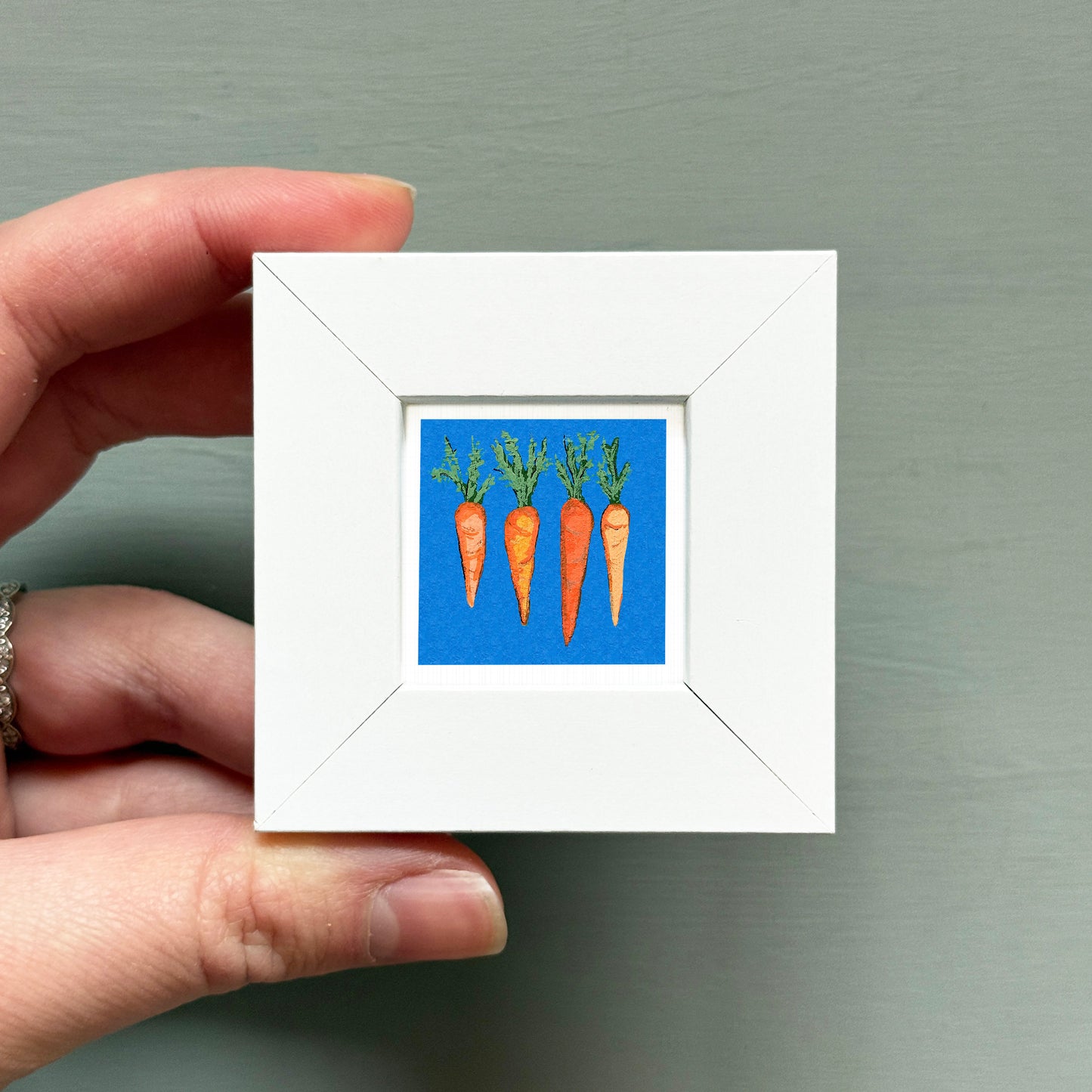 A hand is holding a small white frame containing a painting of three carrots against a blue background.