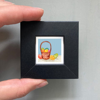 A hand is holding a small black picture frame containing a painting of a basket filled with colorful Easter eggs.
