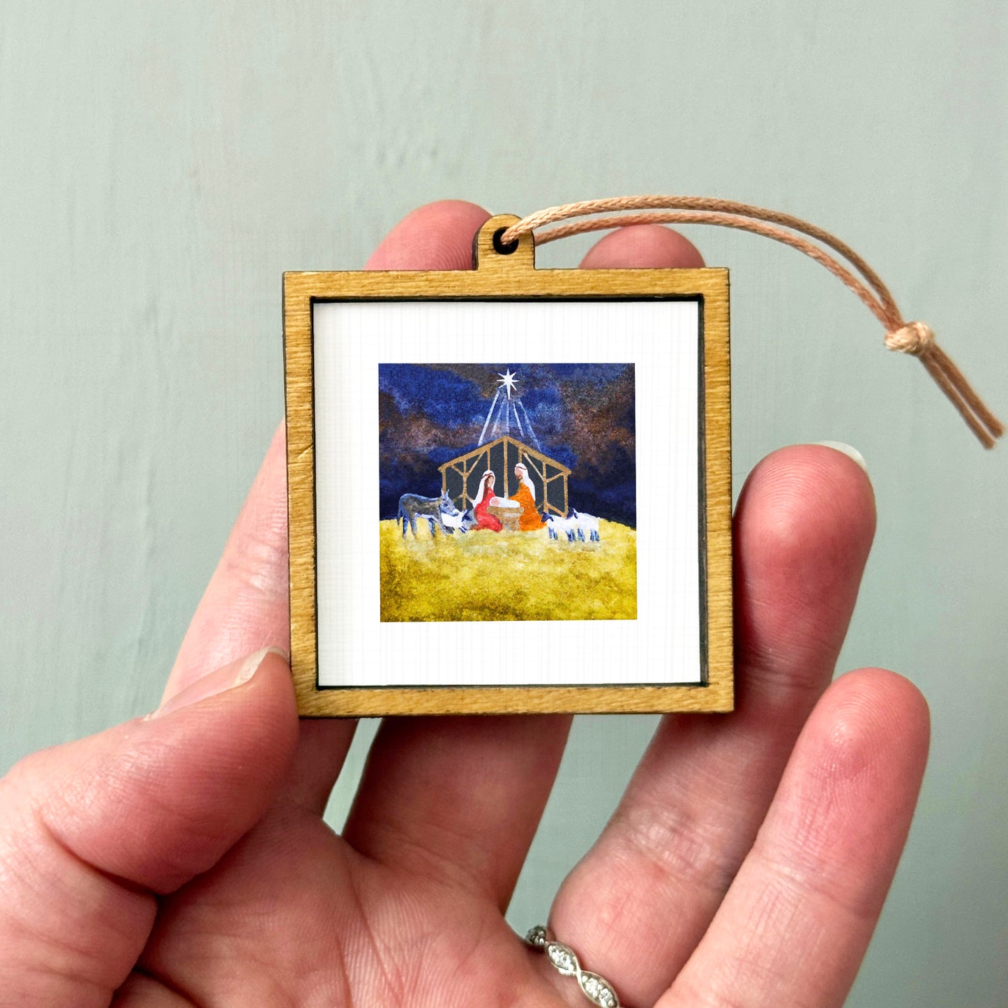 A hand holding a small wooden frame containing a picture of a nativity scene with a man, a baby, and animals in a field.