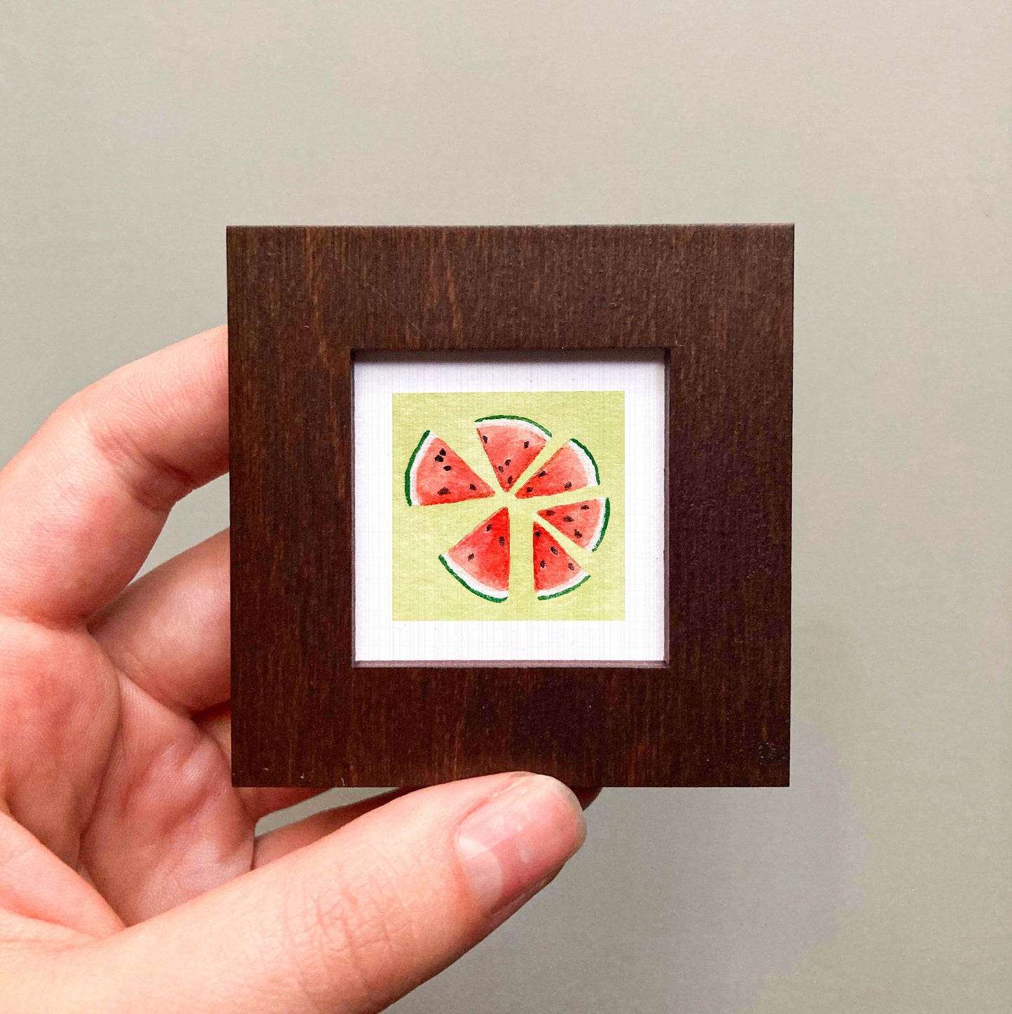 A hand is holding a small wooden frame containing a framed painting of watermelon slices.