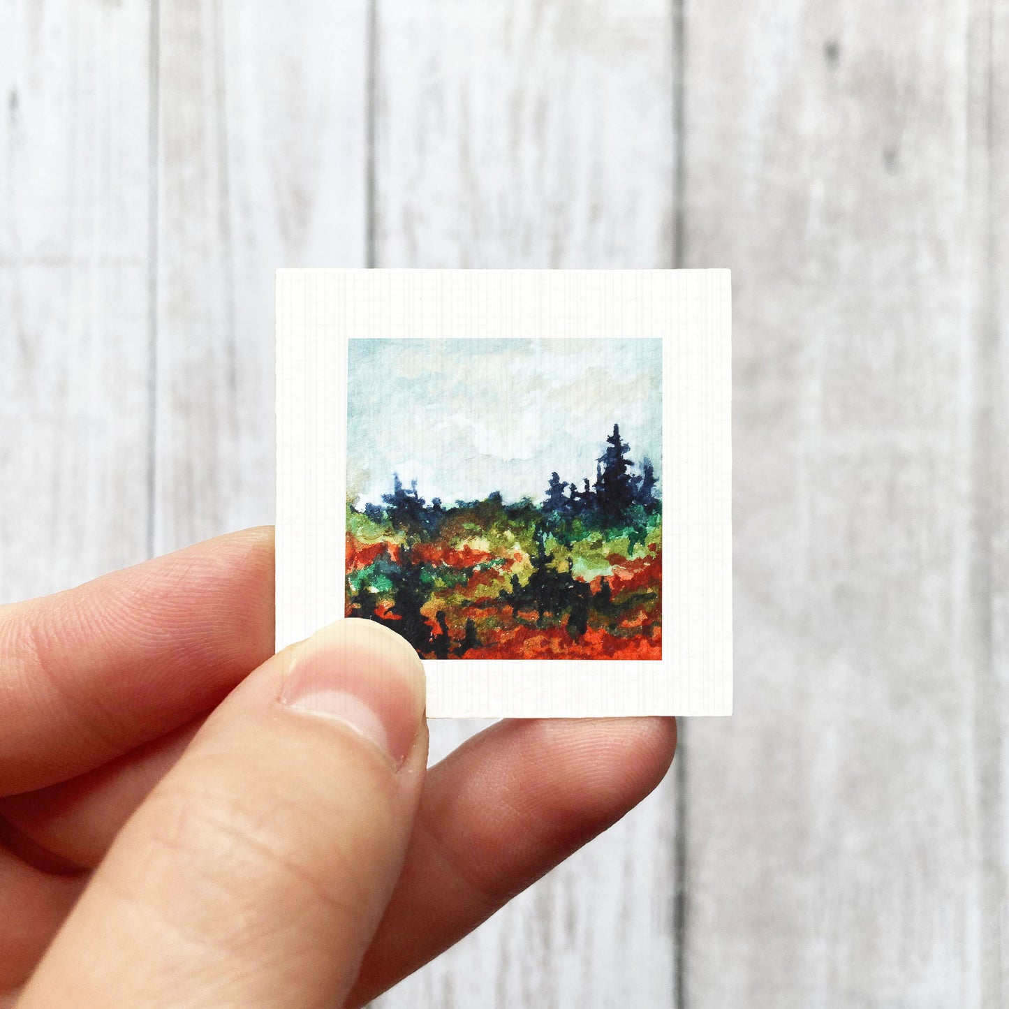 A hand is holding a small square painting of a landscape with trees and a colorful background.