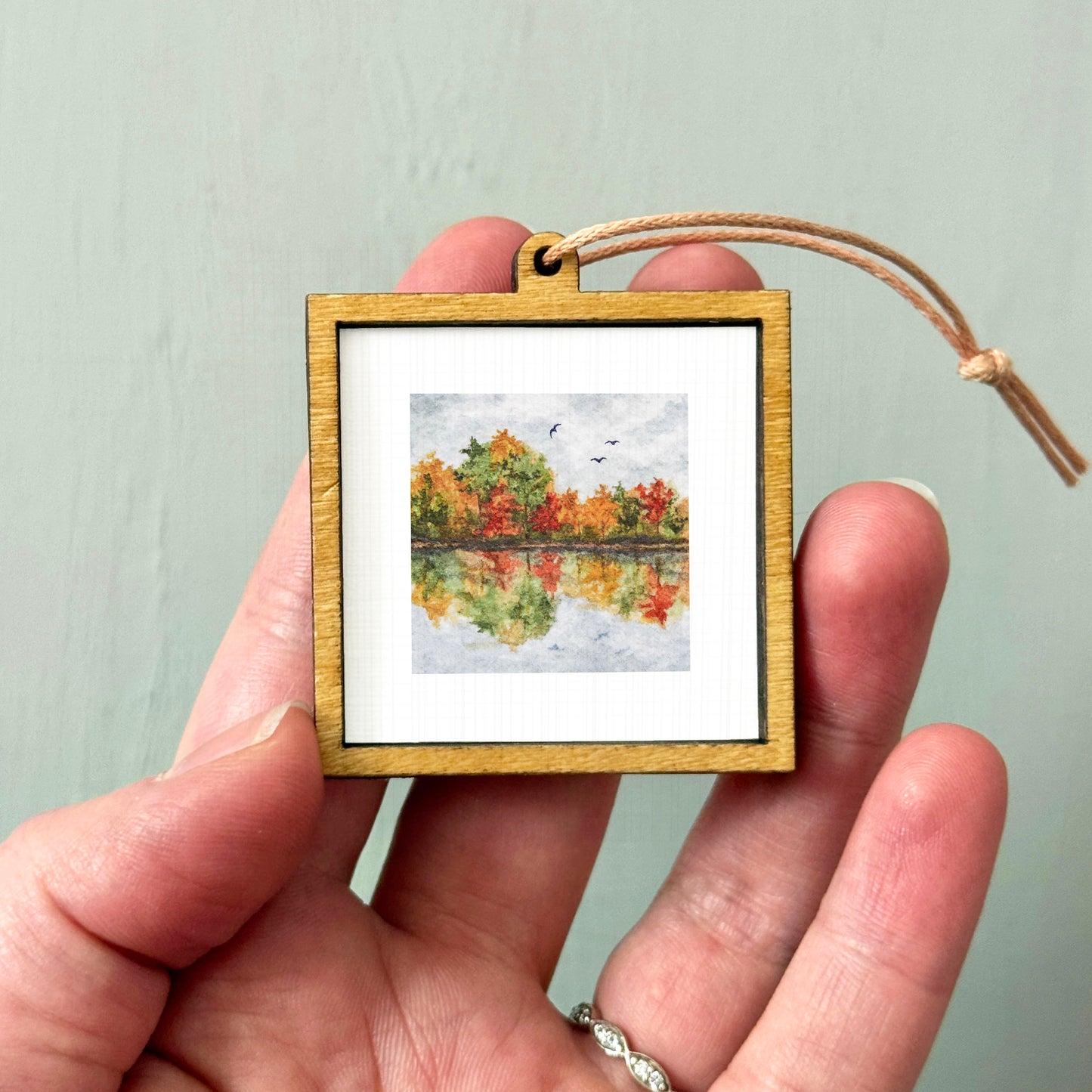 A hand holding a small wooden frame containing a painting of a landscape with trees and a body of water.