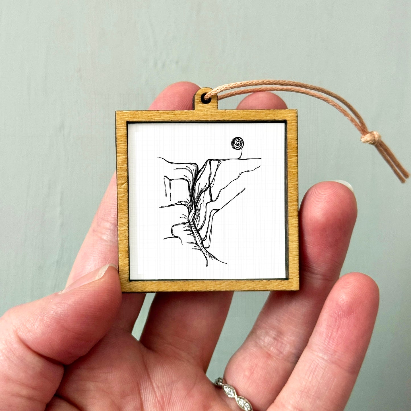 A hand holding a small wooden frame containing a black and white line drawing of a mountain or landscape.