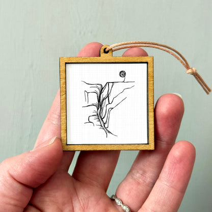 A hand holding a small wooden frame containing a black and white line drawing of a mountain or landscape.