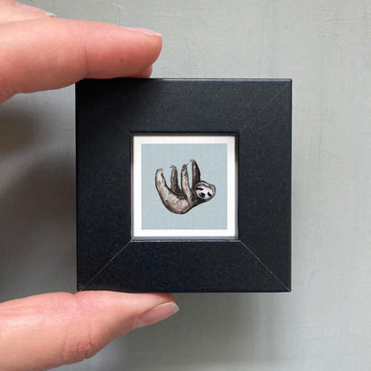 A person's hand is holding a small black frame containing a framed illustration of a sloth.