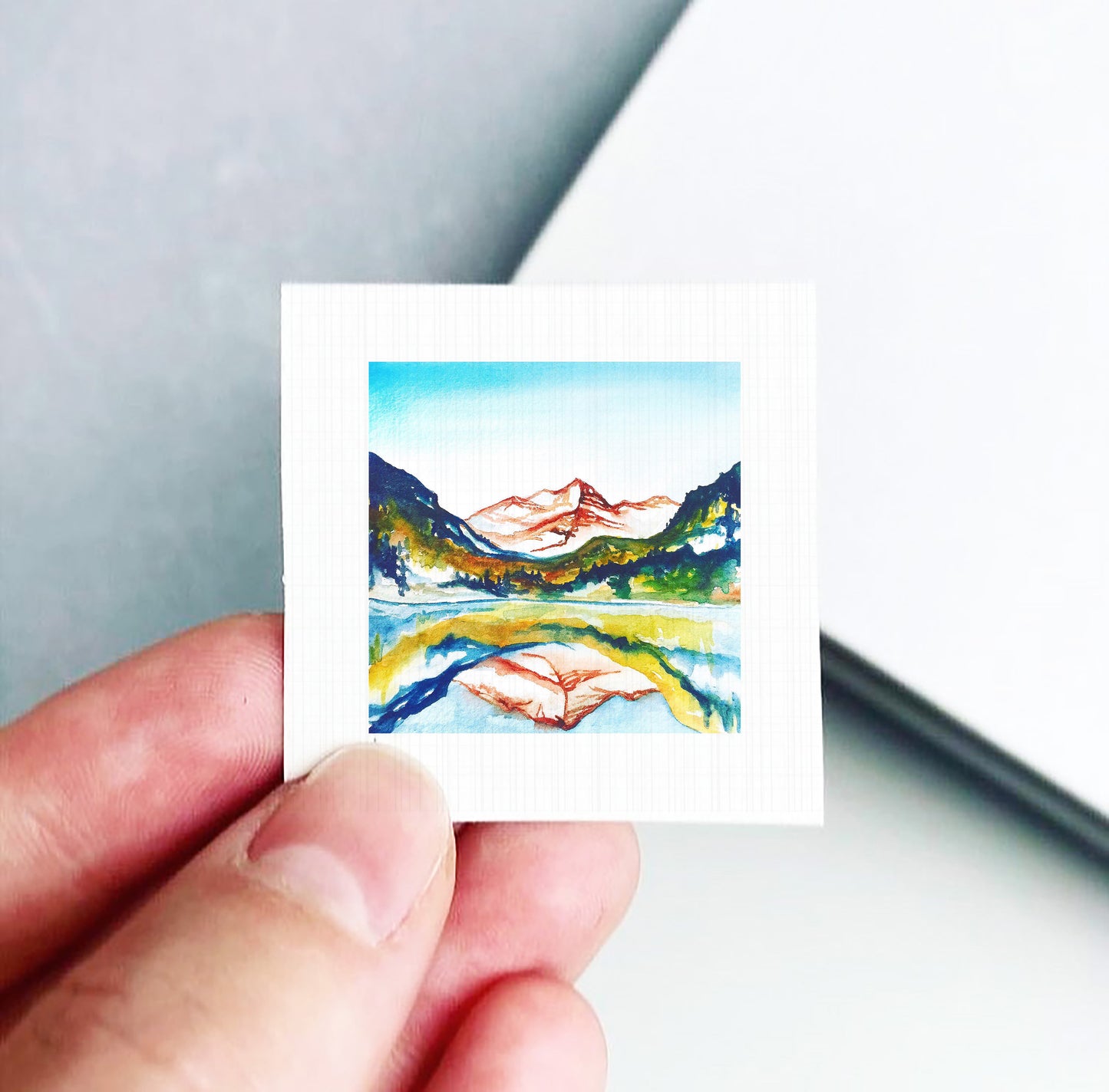 A hand is holding a small square artwork depicting a mountain landscape with a lake and a reflection of the mountains in the water.