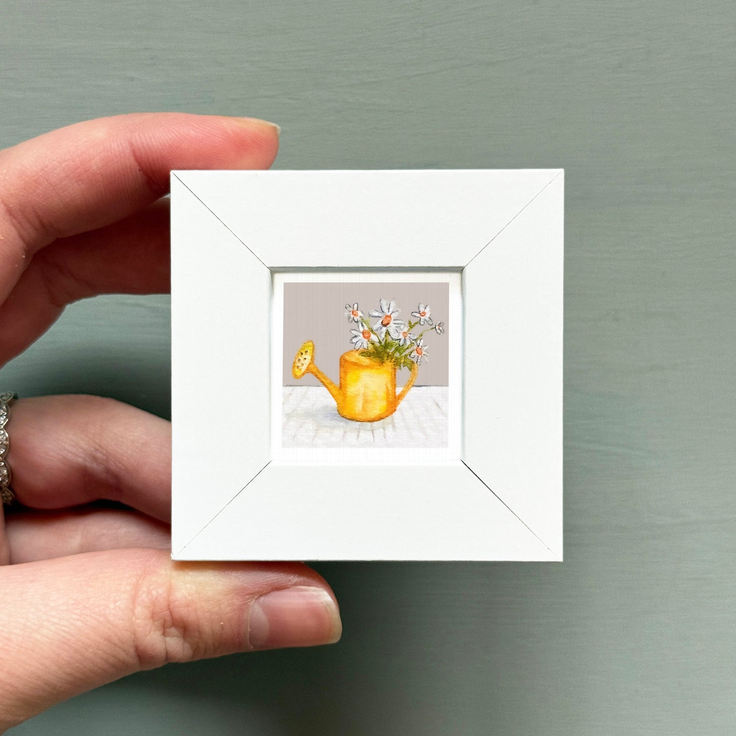 A hand is holding a small white picture frame containing a painting of a yellow watering can with flowers.