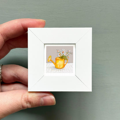 A hand is holding a small white picture frame containing a painting of a yellow watering can with flowers.