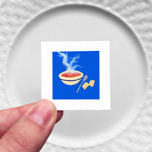 A hand is holding a small square with a vibrant illustration of a bowl of soup, a spoon, and a piece of bread on a blue background.